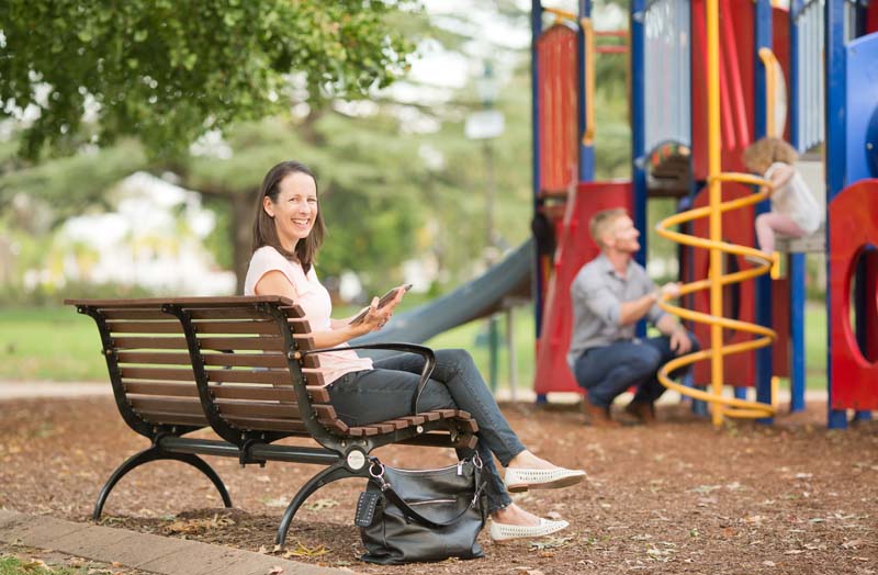 Woman sitting in playground with man and child playing in the background