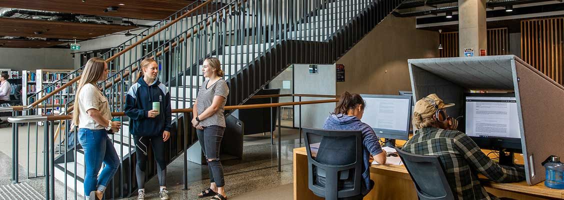 Image taken in library of three students standing near staircase chatting, two students at computers studying