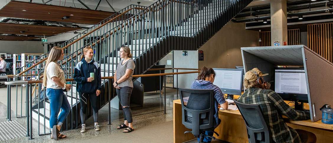 Image taken in library of three students standing near staircase chatting, two students at computers studying