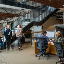 Image taken in library of three students standing near staircase chatting, two students at computers studying